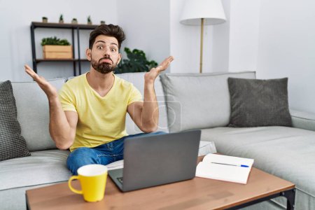 Foto de Young man with beard using laptop at home clueless and confused expression with arms and hands raised. doubt concept. - Imagen libre de derechos