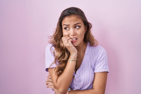 Young hispanic woman standing over pink background looking stressed and nervous with hands on mouth biting nails. anxiety problem. 