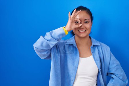 Foto de Asiática joven mujer de pie sobre fondo azul haciendo ok gesto con la mano sonriendo, ojo mirando a través de los dedos con cara feliz. - Imagen libre de derechos