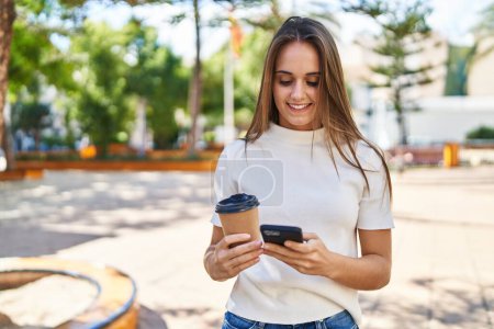 Foto de Mujer rubia joven usando smartphone y tomando café en el parque - Imagen libre de derechos