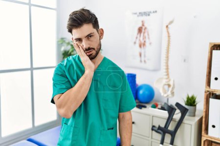Foto de Young man with beard working at pain recovery clinic thinking looking tired and bored with depression problems with crossed arms. - Imagen libre de derechos