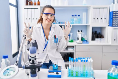Foto de Young hispanic woman working at scientist laboratory showing and pointing up with fingers number eight while smiling confident and happy. - Imagen libre de derechos