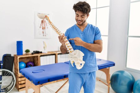 Photo for Young hispanic man wearing physio therapist uniform holding anatomical model of vertebral column at clinic - Royalty Free Image