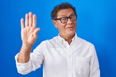 Foto de Middle age hispanic man standing over blue background waiving saying hello happy and smiling, friendly welcome gesture - Imagen libre de derechos