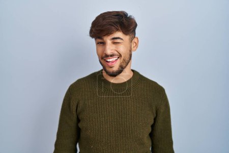 Téléchargez les photos : Arabe homme avec barbe debout sur fond bleu clin d'oeil en regardant la caméra avec une expression sexy, gai et heureux visage. - en image libre de droit