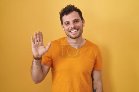 Téléchargez les photos : Jeune homme hispanique debout sur fond jaune renonçant à dire bonjour heureux et souriant, geste de bienvenue amical - en image libre de droit