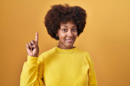 Foto de Joven mujer afroamericana de pie sobre fondo amarillo mostrando y señalando hacia arriba con el dedo número uno mientras sonríe confiado y feliz. - Imagen libre de derechos