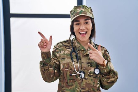 Foto de Joven mujer del sur asiático vistiendo camuflaje ejército uniforme y estetoscopio sonriendo y mirando a la cámara apuntando con dos manos y dedos a un lado. - Imagen libre de derechos