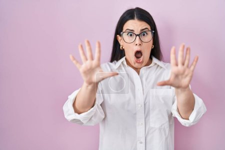 Photo for Young brunette woman standing over pink background afraid and terrified with fear expression stop gesture with hands, shouting in shock. panic concept. - Royalty Free Image
