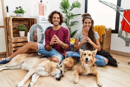 Photo for Young hispanic couple doing laundry with dogs hands together and fingers crossed smiling relaxed and cheerful. success and optimistic - Royalty Free Image