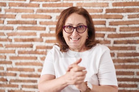 Photo for Senior woman with glasses standing over bricks wall with hands together and crossed fingers smiling relaxed and cheerful. success and optimistic - Royalty Free Image
