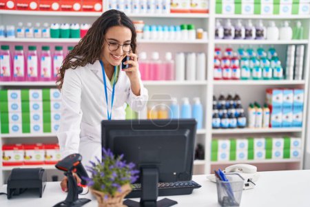 Photo for Young beautiful hispanic woman pharmacist talking on smartphone using computer at pharmacy - Royalty Free Image