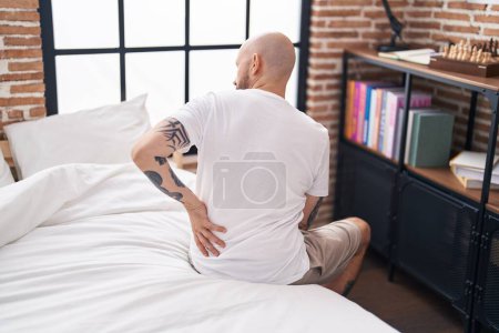 Foto de Joven hombre calvo sufriendo por lesiones en la espalda sentado en la cama en el dormitorio - Imagen libre de derechos