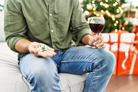 Foto de Young hispanic man holding glass of wine and pills sitting on sofa with dog by christmas tree at home - Imagen libre de derechos