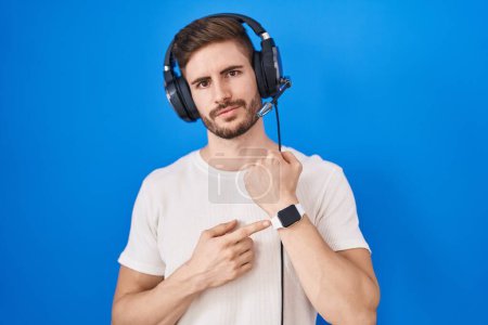 Téléchargez les photos : Homme hispanique avec barbe écoutant de la musique portant des écouteurs pressés pointant vers le temps de regarder, impatience, regardant la caméra avec une expression détendue - en image libre de droit