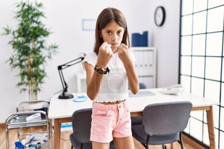Photo for Young hispanic girl standing at pediatrician clinic ready to fight with fist defense gesture, angry and upset face, afraid of problem - Royalty Free Image