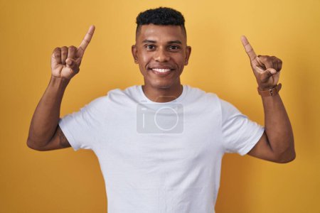 Foto de Young hispanic man standing over yellow background smiling amazed and surprised and pointing up with fingers and raised arms. - Imagen libre de derechos