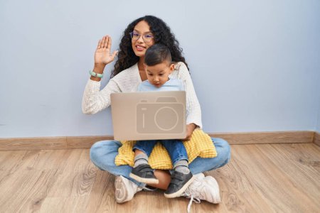 Foto de Joven madre hispana y niño usando computadora portátil sentado en el suelo renunciando a decir hola feliz y sonriente, gesto de bienvenida amistoso - Imagen libre de derechos