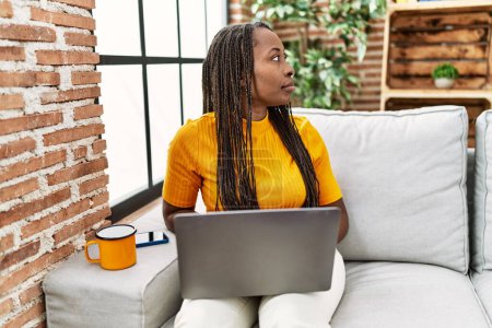 Foto de Mujer africana sentada en el sofá utilizando el ordenador portátil en casa mirando a un lado, relajarse pose perfil con la cara natural con sonrisa confiada. - Imagen libre de derechos