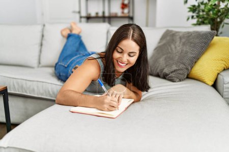 Téléchargez les photos : Jeune belle femme hispanique écrivant sur un cahier couché sur un canapé à la maison - en image libre de droit