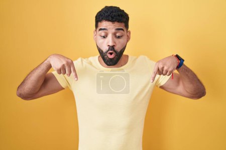 Foto de Hispanic man with beard standing over yellow background pointing down with fingers showing advertisement, surprised face and open mouth - Imagen libre de derechos