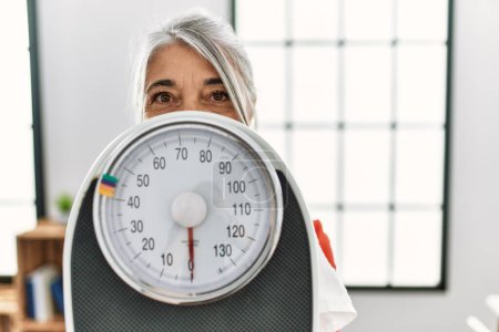 Téléchargez les photos : Middle age grey-haired woman covering face with weighing machine at home - en image libre de droit