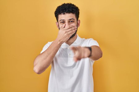 Téléchargez les photos : Arabe debout sur fond jaune se moquant de vous, pointant du doigt la caméra avec la main sur la bouche, expression de honte - en image libre de droit