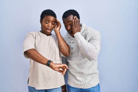 Photo for Young african american couple standing over blue background together looking at the watch time worried, afraid of getting late - Royalty Free Image
