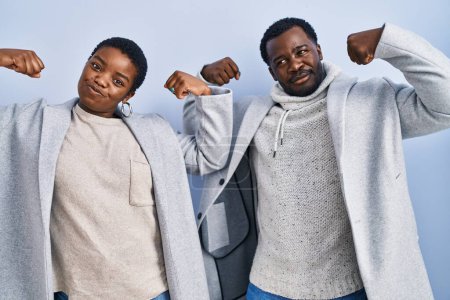 Foto de Joven pareja afroamericana de pie sobre fondo azul juntos mostrando los músculos de los brazos sonriendo orgulloso. concepto de fitness. - Imagen libre de derechos