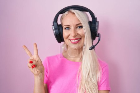 Téléchargez les photos : Femme caucasienne écoutant de la musique à l'aide d'écouteurs souriant avec un clin d'œil heureux à la caméra faisant signe de victoire. numéro deux. - en image libre de droit