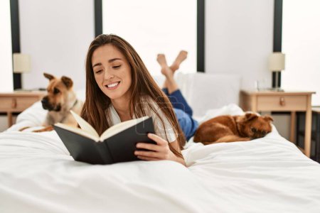 Téléchargez les photos : Jeune femme hispanique lecture livre couché sur le lit avec des chiens dans la chambre - en image libre de droit