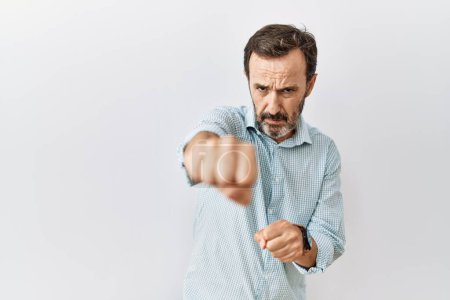 Foto de Hombre hispano de mediana edad con barba de pie sobre fondo aislado golpeando puño para luchar, ataque agresivo y enojado, amenaza y violencia - Imagen libre de derechos