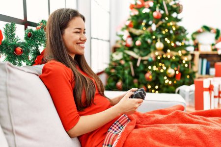 Photo for Young latin woman playing video game sitting by christmas tree at home - Royalty Free Image