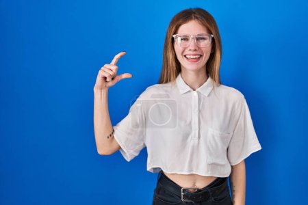 Photo for Beautiful woman standing over blue background smiling and confident gesturing with hand doing small size sign with fingers looking and the camera. measure concept. - Royalty Free Image
