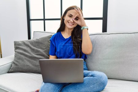 Foto de Mujer morena joven usando el ordenador portátil en casa haciendo buen gesto con la mano sonriendo, los ojos mirando a través de los dedos con la cara feliz. - Imagen libre de derechos