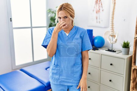 Photo for Beautiful blonde physiotherapist woman working at pain recovery clinic smelling something stinky and disgusting, intolerable smell, holding breath with fingers on nose. bad smell - Royalty Free Image