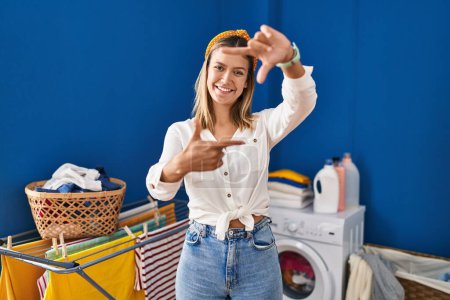 Photo for Young blonde woman at laundry room smiling making frame with hands and fingers with happy face. creativity and photography concept. - Royalty Free Image