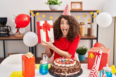 Téléchargez les photos : Femme hispanique avec les cheveux bouclés célébrant anniversaire avec gâteau et présent célébrant fou et étonné pour le succès avec les yeux ouverts hurlant excité. - en image libre de droit