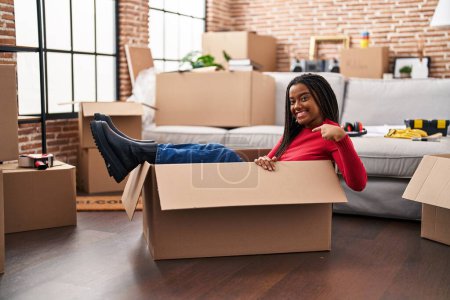 Téléchargez les photos : Jeune afro-américain avec des tresses se déplaçant vers une nouvelle maison à l'intérieur d'une boîte en carton pointant du doigt un sourire heureux et fier - en image libre de droit