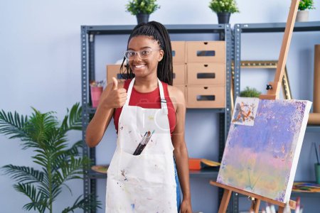 Foto de Young african american with braids at art studio painting on canvas smiling happy and positive, thumb up doing excellent and approval sign - Imagen libre de derechos