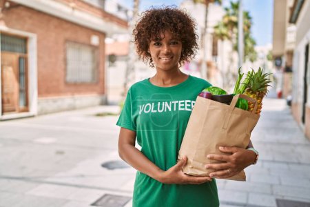 Photo for Young african american woman wearing volunteer uniform holding groceries at street - Royalty Free Image