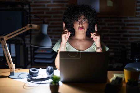 Photo for Young brunette woman with curly hair working at the office at night pointing up looking sad and upset, indicating direction with fingers, unhappy and depressed. - Royalty Free Image