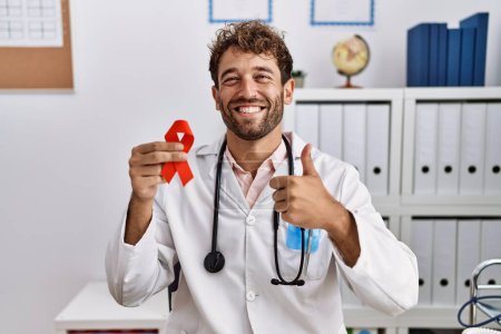 Photo for Young hispanic doctor man holding support red ribbon at medical clinic smiling happy and positive, thumb up doing excellent and approval sign - Royalty Free Image