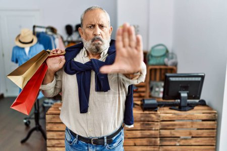 Photo for Handsome senior man holding shopping bags at boutique shop doing stop sing with palm of the hand. warning expression with negative and serious gesture on the face. - Royalty Free Image