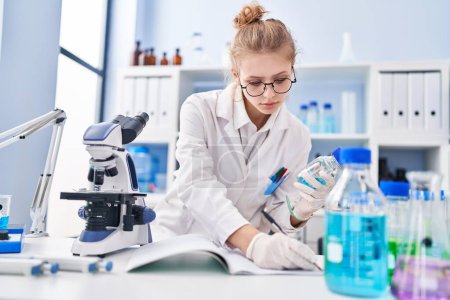 Téléchargez les photos : Young blonde woman scientist writing on notebook holding bottle at laboratory - en image libre de droit