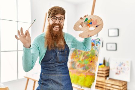 Téléchargez les photos : Jeune rousse homme souriant confiant tenant pinceau et palette au studio d'art - en image libre de droit