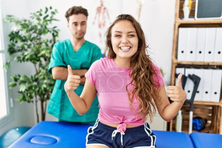 Photo for Young hispanic woman at physiotherapist appointment success sign doing positive gesture with hand, thumbs up smiling and happy. cheerful expression and winner gesture. - Royalty Free Image