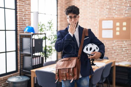 Photo for Hispanic man with beard holding bike helmet at the office covering mouth with hand, shocked and afraid for mistake. surprised expression - Royalty Free Image