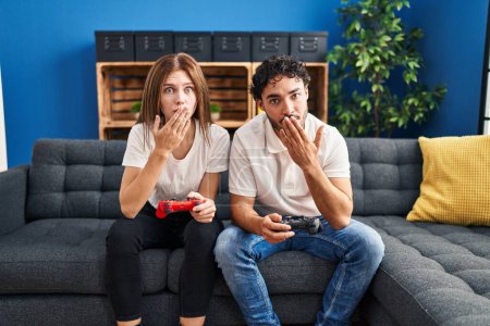 Photo for Young couple playing video game holding controller at home covering mouth with hand, shocked and afraid for mistake. surprised expression - Royalty Free Image