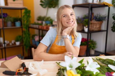 Photo for Young blonde woman florist smiling confident sitting on table at flower shop - Royalty Free Image